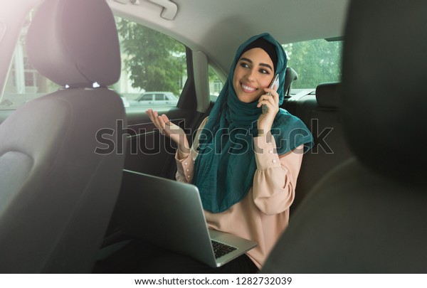 Smiling arabian businesswomen with\
hijab talking by phone using laptop on the back seat in\
car.