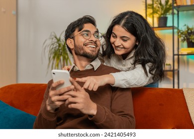 Smiling Arabian woman talking with boyfriend swiping smartphone sitting on sofa at home. Multiethnic young couple using mobile phone together making order online delivery in living room apartment. - Powered by Shutterstock