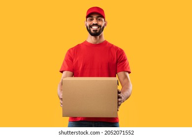 Smiling Arabic Delivery Guy Holding Parcel Packed In Cardboard Box Standing Smiling To Camera Posing Over Yellow Background In Studio, Wearing Red Uniform. Transportation And Delivering Service