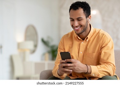 Smiling Arabic Male Listening To Music On Mobile Phone Wearing Wireless Headphones Sitting On Couch Relaxing At Home. Male Enjoying Favorite Playlist Using Musical Application, Free Copy Space