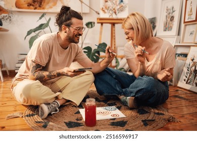 Smiling artistic fun couple sitting on a floor at creative art studio and acting childish. Young hipster painter couple painting on each other face and having fun together at creative atelier. - Powered by Shutterstock