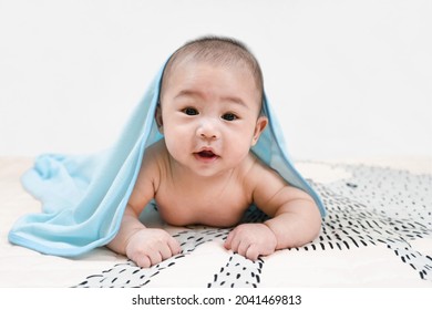 Smiling Asian Baby Boy Looking At Camera Under A Blue Towel Crawling On Bed. Happy Kid Portrait. Lying At Home Bed. Infant Playing. Face Skin Care. Good Emotion.