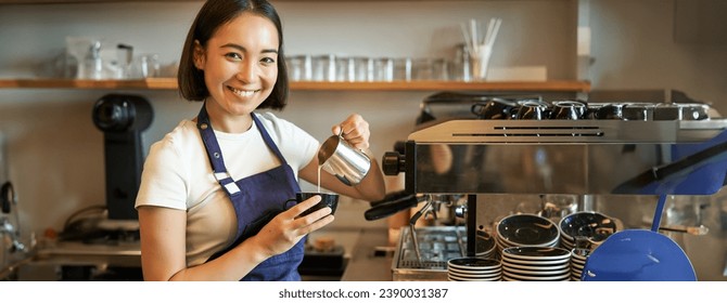 Smiling asian barista girl, making coffee, pouring steamed milk into cappuccino, doing latte art in cup, working in cafe. - Powered by Shutterstock