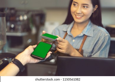 Smiling Asian Barista Using Smartphone And Nfs Technology To Help Customer Pay For A Purchase With Phone, Mobile Payment, Online Shop At Coffee Cafe.
