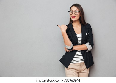 Smiling asian business woman in eyeglasses pointing and looking away over gray background