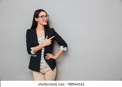 Smiling Asian Business Woman In Eyeglasses With Arm On Hip Pointing And Looking Away Over Gray Background