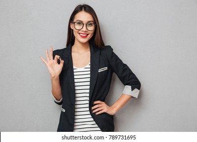 Smiling asian business woman in eyeglasses with arm on hip showing ok sign and looking at the camera over gray background