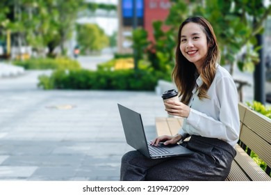 Smiling Asian Business Woman Wearing Trendy Sitting In The Central City Street And Uses Her Phone And Laptop. Pretty Summer Woman In White Shirt Walks Down The Street Looking At Her Mobile Phone