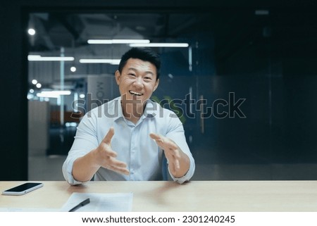 Smiling asian businessman talking on video call, looking at camera while sitting at desk in office. Gestures, discusses, explains.