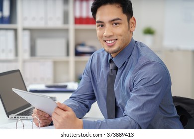 Smiling Asian businessman working in an office seated at a desk with a tablet in his hand smiling at the camera