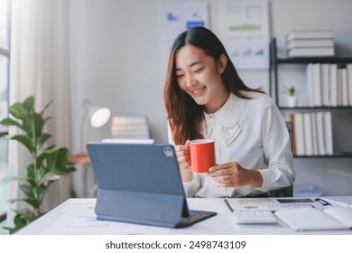 Smiling asian businesswoman drinking coffee while working from her home office, using a tablet to check financial charts - Powered by Shutterstock