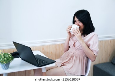 Smiling Asian businesswoman is drinking and relaxing with a cup of coffee and online working with laptop for work from home. Lifstyle of businesspeople with techonology concept.