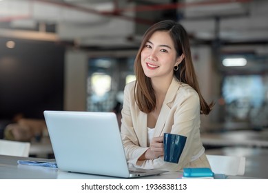 Smiling Asian businesswoman holding a coffee mug and laptop at the office. Looking at the camera.
