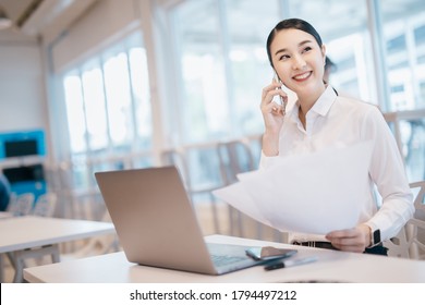 Smiling Asian Businesswoman Talking With Partner While Sitting At Her Desk. Professional Employees Discussing Ideas Of Project On Laptop.
