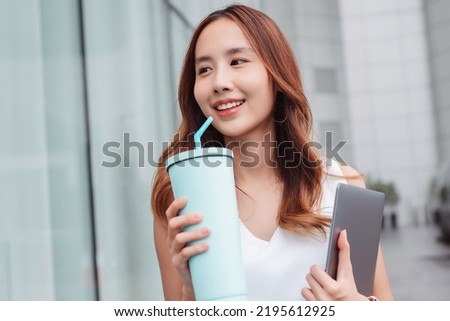 Smiling asian businesswoman use smartphone and hold reusable eco-friendly ecological cup and laptop computer while commuting in city in the morning
