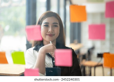 Smiling Asian businesswoman using colorful sticky notes for brainstorming on a glass wall in a modern office.

 - Powered by Shutterstock