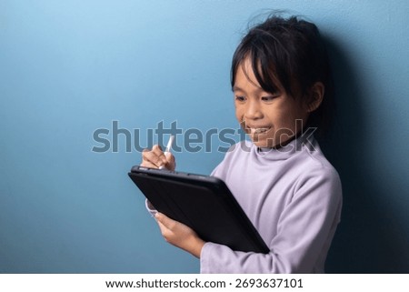 Smiling Asian child using digital tablet with stylus for online learning on blue wall background, concept of happy education, creative learning, and digital technology for kids.