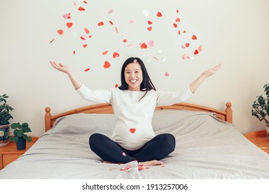 Smiling Asian Chinese Pregnant Woman Sitting On Bed Throwing Small Red Pink Paper Hearts. Expecting Excited Young Lady Female At Home. Healthy Happy Parenthood Lifestyle. Love Is In The Air.