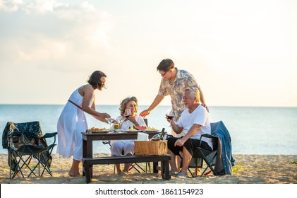 Smiling Asian Couple Man And Woman With Retired Senior Parents Enjoy Dinner Party Together On Island Beach At Summer Sunset. Happy Two Generation Family Relax And Having Fun With On Holiday Vacation