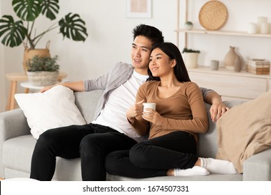 Smiling Asian Couple Relaxing And Watching TV At Home, Having Rest After Hard Moving Day, Copy Space