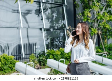 Smiling Asian Curly Business Woman Wearing Trendy Walks Down The Central City Street And Uses Her Phone. Pretty Summer Woman In White Shirt Walks Down The Street Looking At Her Mobile Phone