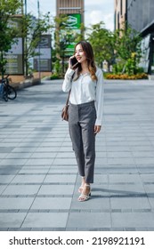 Smiling Asian Curly Business Woman Wearing Trendy Walks Down The Central City Street And Uses Her Phone. Pretty Summer Woman In White Shirt Walks Down The Street Looking At Her Mobile Phone