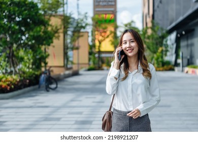 Smiling Asian Curly Business Woman Wearing Trendy Walks Down The Central City Street And Uses Her Phone. Pretty Summer Woman In White Shirt Walks Down The Street Looking At Her Mobile Phone