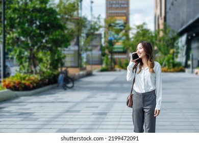 Smiling Asian Curly Business Woman Wearing Trendy Walks Down The Central City Street And Uses Her Phone. Pretty Summer Woman In White Shirt Walks Down The Street Looking At Her Mobile Phone