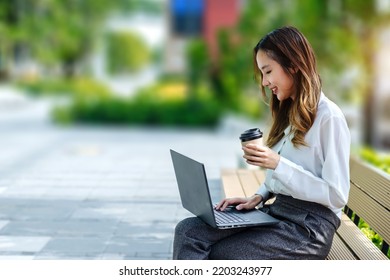 Smiling Asian Curly Business Woman Wearing Trendy Sitting In The Central City Street And Uses Her Phone And Laptop. Pretty Summer Woman In White Shirt Walks Down The Street Looking At Her Mobile Phone