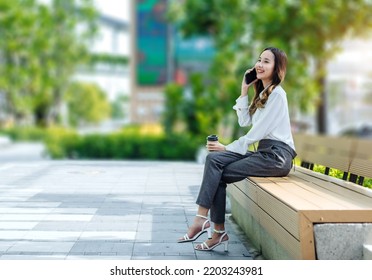 Smiling Asian Curly Business Woman Wearing Trendy Walks Down The Central City Street And Uses Her Phone. Pretty Summer Woman In White Shirt Walks Down The Street Looking At Her Mobile Phone