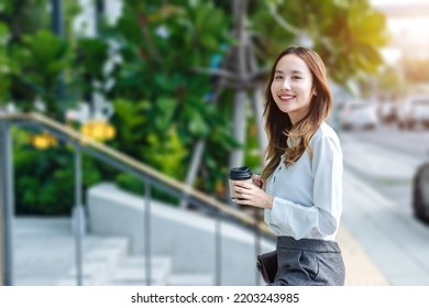 Smiling Asian Curly Business Woman Wearing Trendy Walks Down The Central City Street And Uses Her Phone. Pretty Summer Woman In White Shirt Walks Down The Street Looking At Her Mobile Phone