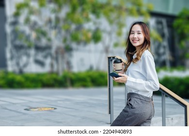 Smiling Asian Curly Business Woman Wearing Trendy Walks Down The Central City Street And Uses Her Phone. Pretty Summer Woman In White Shirt Walks Down The Street Looking At Her Mobile Phone