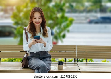 Smiling Asian Curly Business Woman Wearing Trendy Walks Down The Central City Street And Uses Her Phone. Pretty Summer Woman In White Shirt Walks Down The Street Looking At Her Mobile Phone