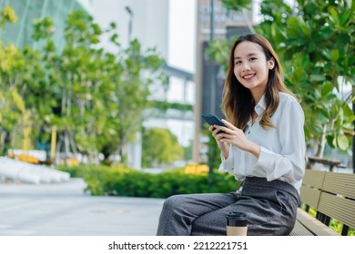 Smiling Asian Curly Business Woman Wearing Trendy Walks Down The Central City Street And Uses Her Phone. Pretty Summer Woman In White Shirt Walks Down The Street Looking At Her Mobile Phone