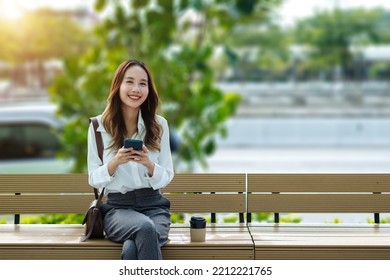 Smiling Asian Curly Business Woman Wearing Trendy Walks Down The Central City Street And Uses Her Phone. Pretty Summer Woman In White Shirt Walks Down The Street Looking At Her Mobile Phone