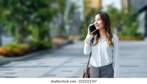 Smiling Asian Curly Business Woman Wearing Trendy Walks Down The Central City Street And Uses Her Phone. Pretty Summer Woman In White Shirt Walks Down The Street Looking At Her Mobile Phone