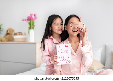 Smiling Asian Cute Teen Girl In Pajama Closes Eyes To Millennial Woman Hold Postcard In Bedroom Interior, Daughter Congratulate Mom. Surprise At Mothers Day, Celebration At Home Due Covid-19 Outbreak