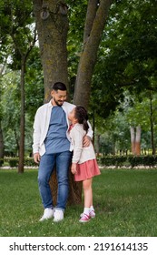 Smiling Asian Dad Hugging And Looking At Daughter Near Tree In Park