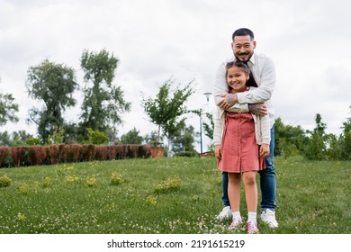 Smiling Asian Dad Hugging Preteen Daughter In Park