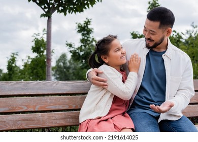 Smiling Asian Dad Hugging And Talking To Daughter On Bench In Park