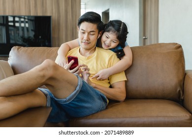 Smiling Asian Daughter Embracing Father From Behind While Looking At Smartphone. Asian Family Sharing Social Media On Mobile Phone, Happy Father Sitting On The Couch In The Living Room At Home