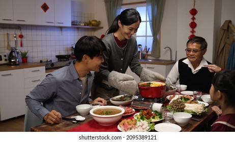 Smiling Asian Daughter In Law Carrying And Putting Hot Pot On Portable Stove. Happy Senior Father Gesturing To Sit Down At Dining Table, Ready To Start Reunion Meal