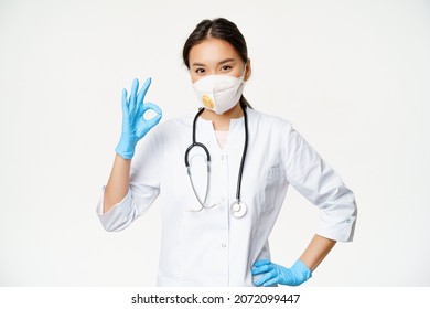 Smiling Asian Doctor, Female Physician Shows Okay Sign, Wears Respirator From Catching Covid Disease, Standing In Uniform Over White Background