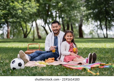 Smiling Asian Family Holding Orange Juice Near Food And Football On Blanket In Park