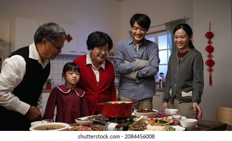 Smiling Asian Father Serving Hot Soup On The Dining Table And Posing With Folded Arms, Feeling Proud Of His Cooking. Preparing Food For Family On Chinese New Year's Eve. Chinese Text Translation: Luck