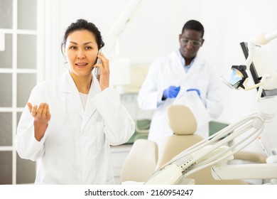 Smiling Asian Female Dentist In White Coat Talking On Mobile Phone In Modern Dental Office..