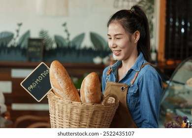 Smiling Asian female holds a basket of bread, wearing a denim shirt and apron in a bakery setting.  happiness of opening a coffee shop in the morning small family business - Powered by Shutterstock