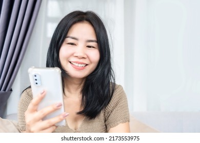 smiling Asian female sitting on sofa using mobile phone enjoin surfing the internet on the weekend - Shutterstock ID 2173553975