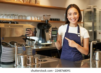 Smiling asian girl barista, cafe owner in apron, showing card machine, payment reader, taking contactless orders in her coffee shop. - Powered by Shutterstock