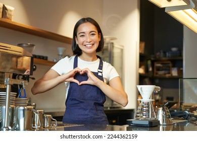Smiling asian girl barista, shows heart sign, loves making coffee an serving clients, standing in uniform behind counter, work in cafe.